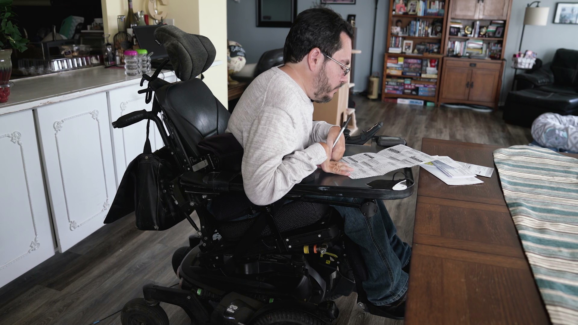 William Crowley fills out a ballot placed on the tray of his wheelchair inside an apartment with kitchen cupboards and a dining table, a chair, bookshelves and other items in the background.