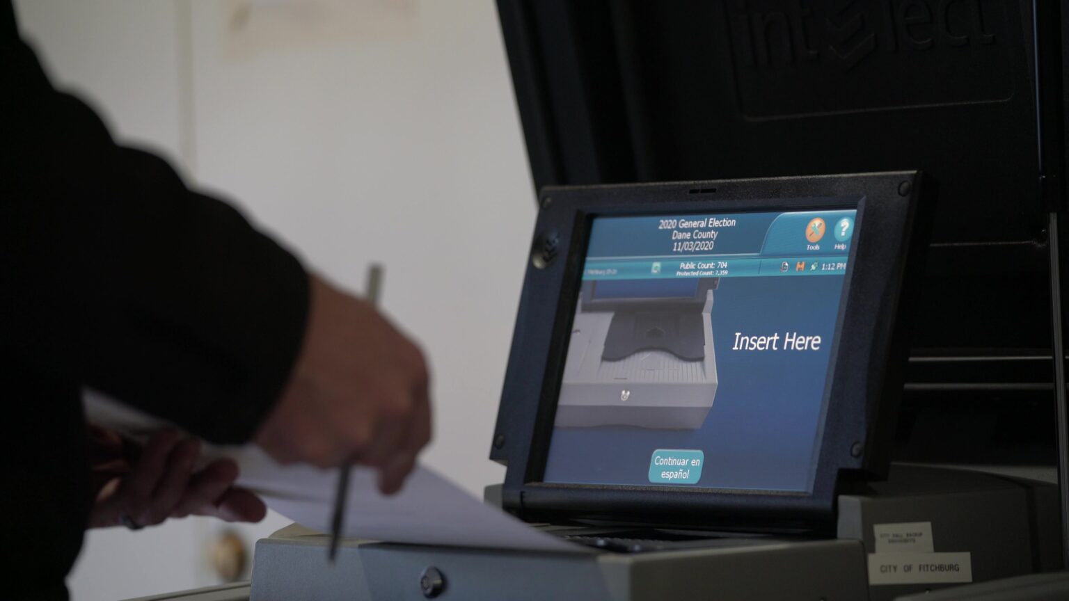 A voter places a ballot into a voting scanner and tabulation machine with a screen that shows the insertion tray with the words Insert Here.