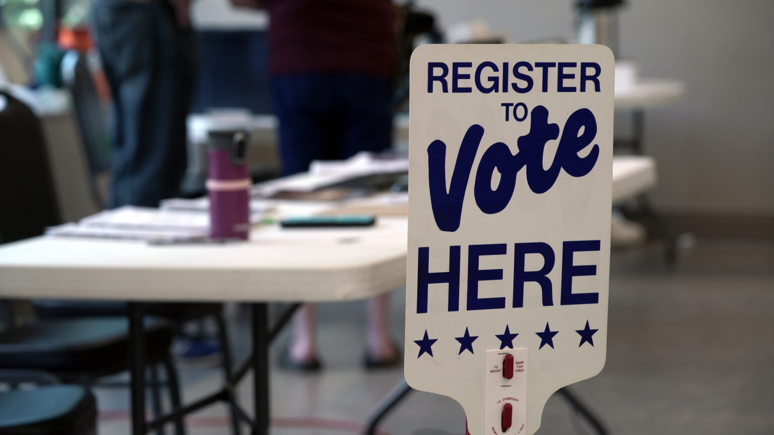 A sign reading Register to Vote Here stands in front of a folding table and chairs, with people standing in the background.