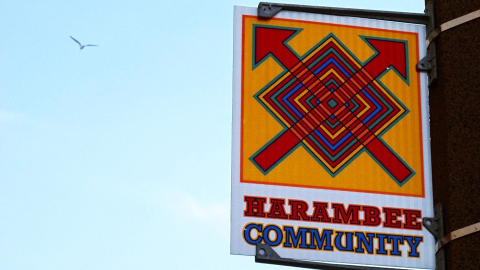A sign mounted to a vertical pole features an abstract graphic with concentric squares and two arrows pointing toward its top corners and the words "Harambee Community."