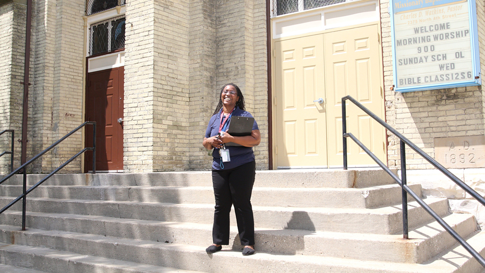 Freda Wright stands on concrete steps in front of the entrance of a brick church with multiple doors, a sign noting times for worship and Sunday school, and a cornerstone marked "A.D. 1892."