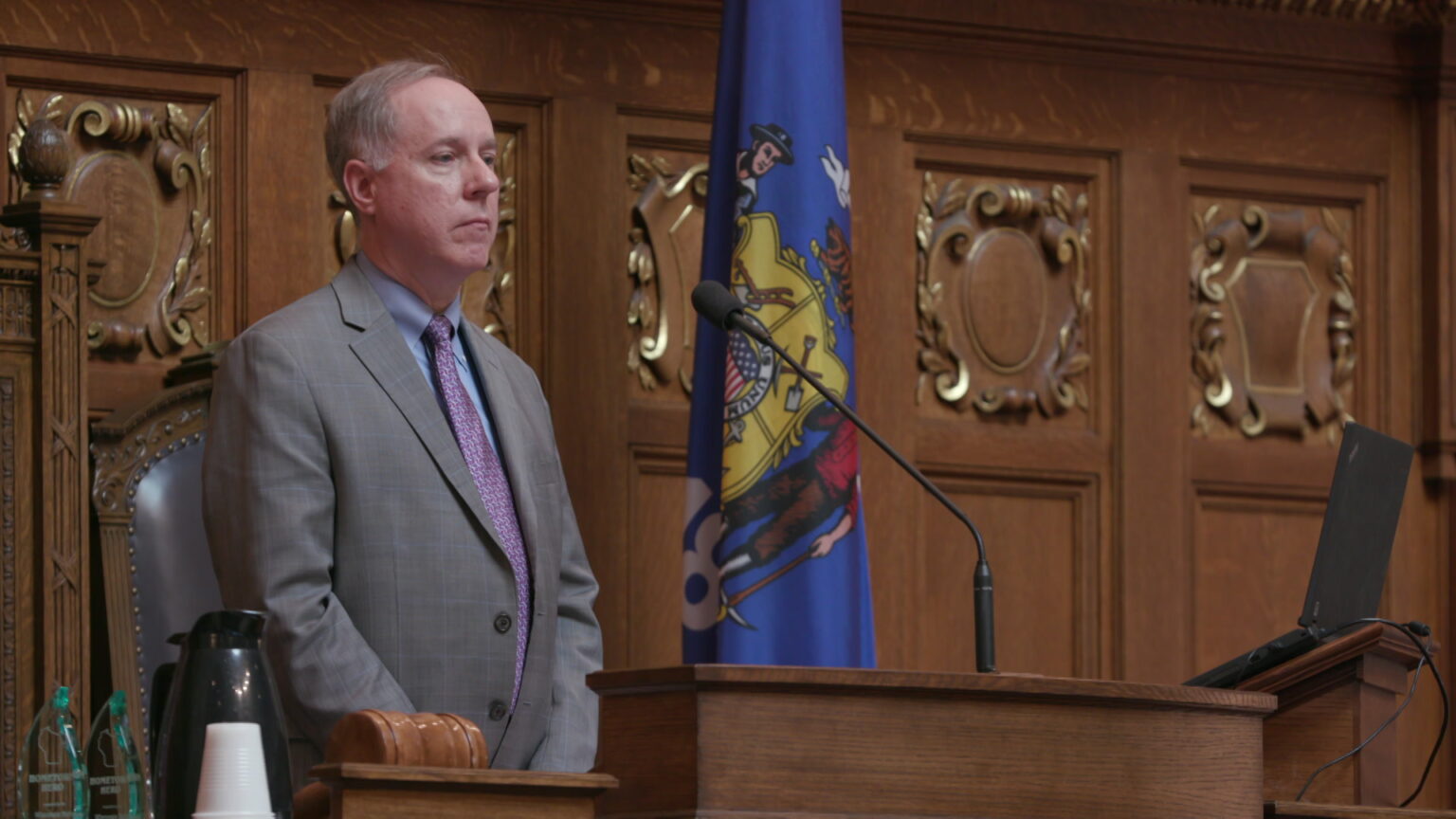 Robin Vos stands from the Speaker's chairs on the dais at the front of the Wisconsin Assembly chamber, with a Wisconsin flag and wood-paneled walls in the background.