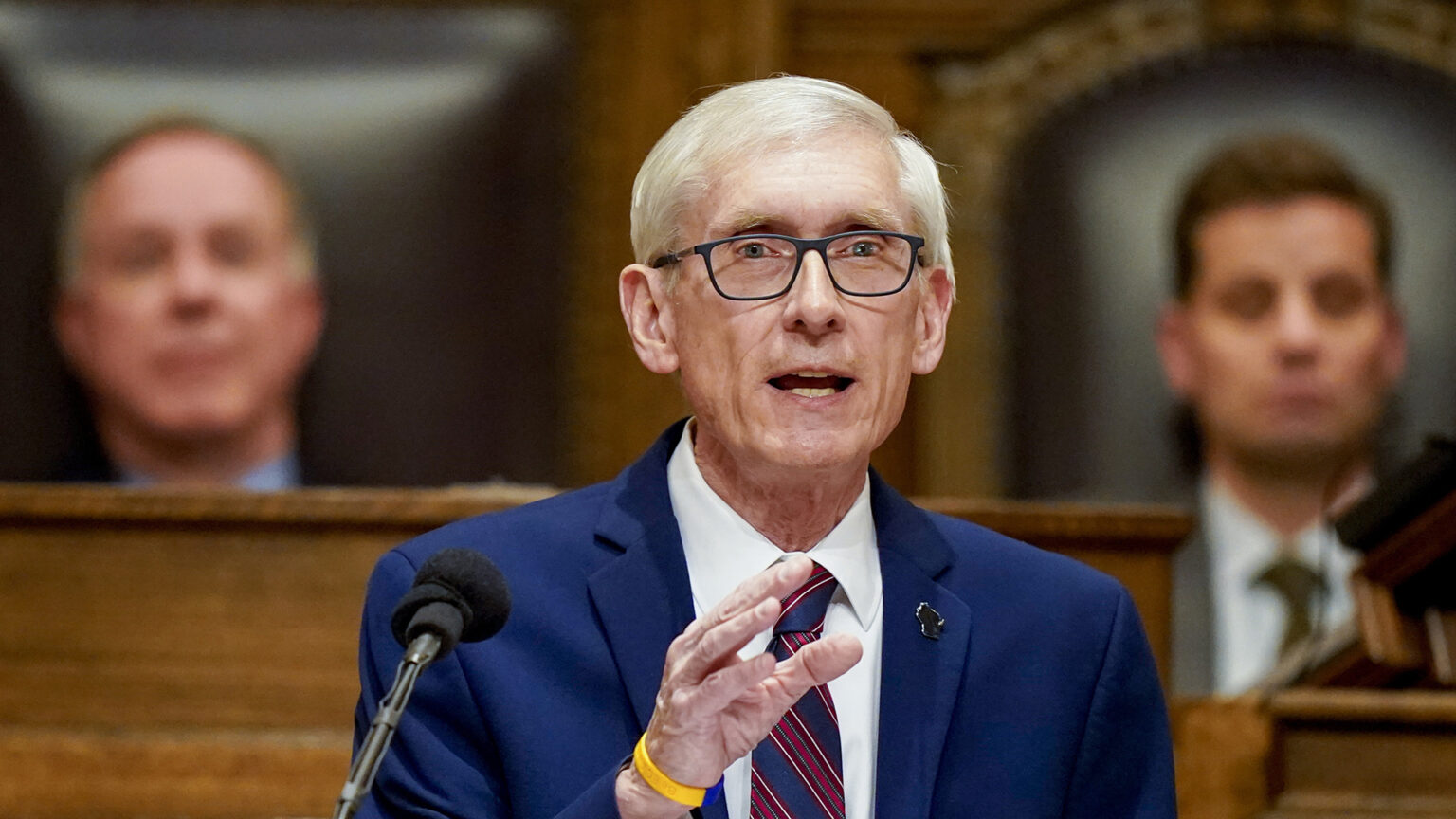 Tony Evers stands at a dais in the Wisconsin Assembly chambers and speaks into a microphone while gesturing with his right hand, with two legislative leadership members seated in the background.