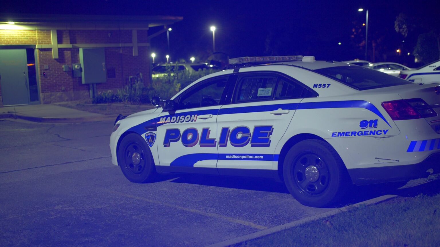 A Madison Police Department cruiser sits in a parking lot with a building and other parked cars in the background, with a blue tint to the image due to a flashing light.