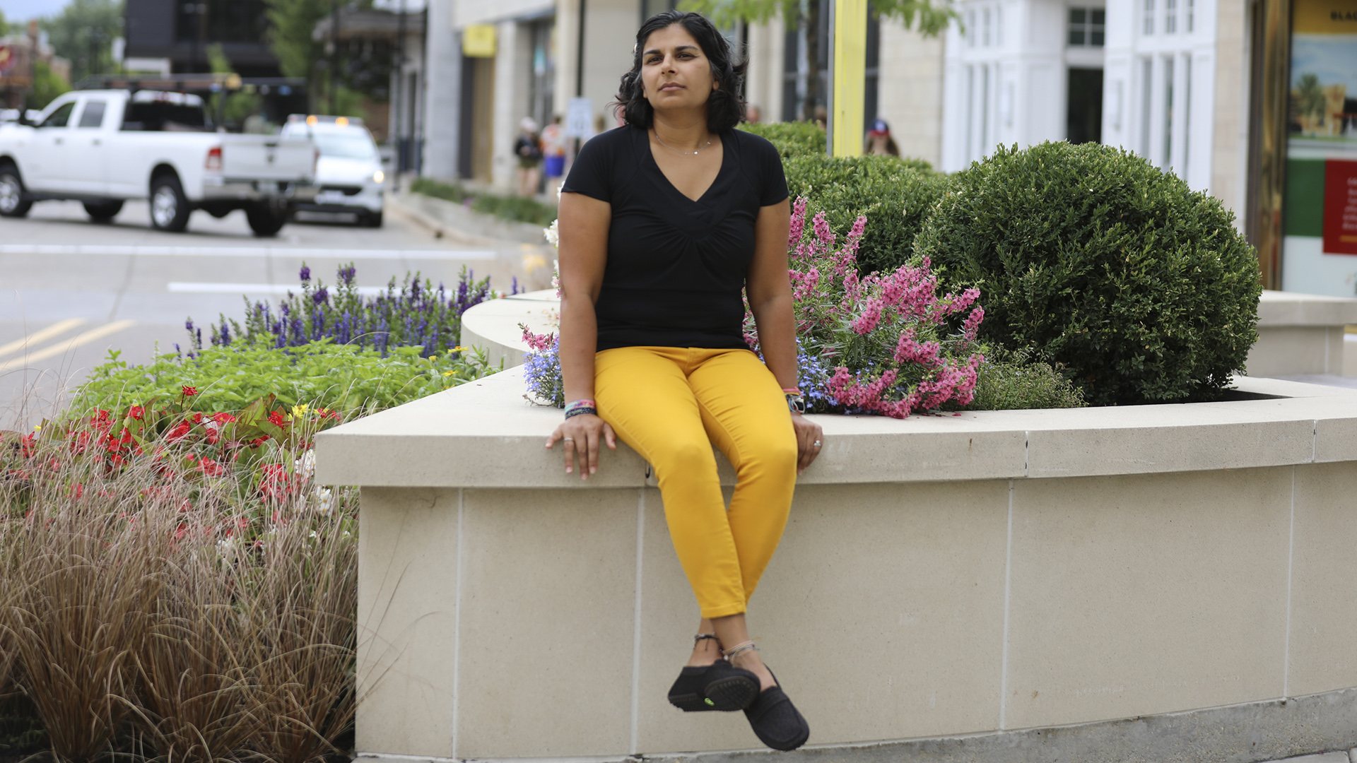 Shefaali Sharma sits on a concrete planter in front of a variety of bushes and shrubs, with a street with cars, pedestrians and building entrances in the background.