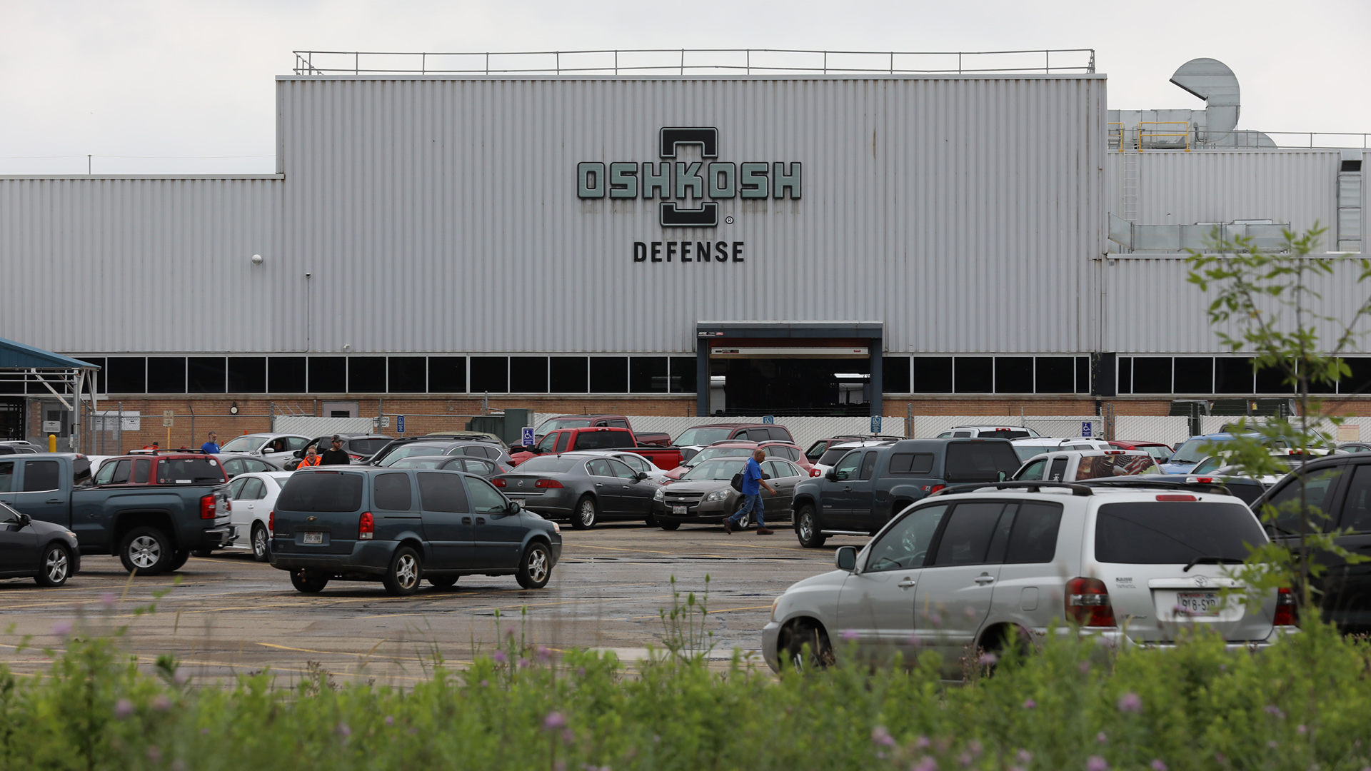 Workers walk in a car-filled parking lot in front of a multi-story brick and corrugated metal building fronted with a large Oshkosh Defense wordmark.