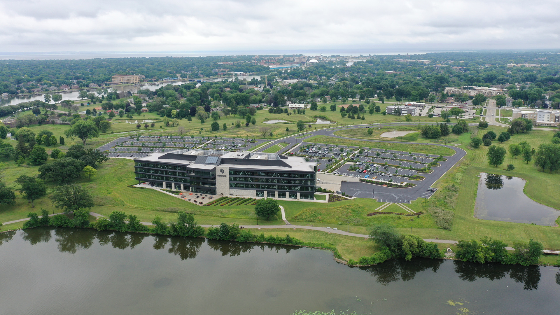 An aerial photo shows a multi-story office building and large adjacent parking lots standing in the midst of fields with green turf and numerous trees, with a lakeshore in the foreground and a forested urban landscape in the background.