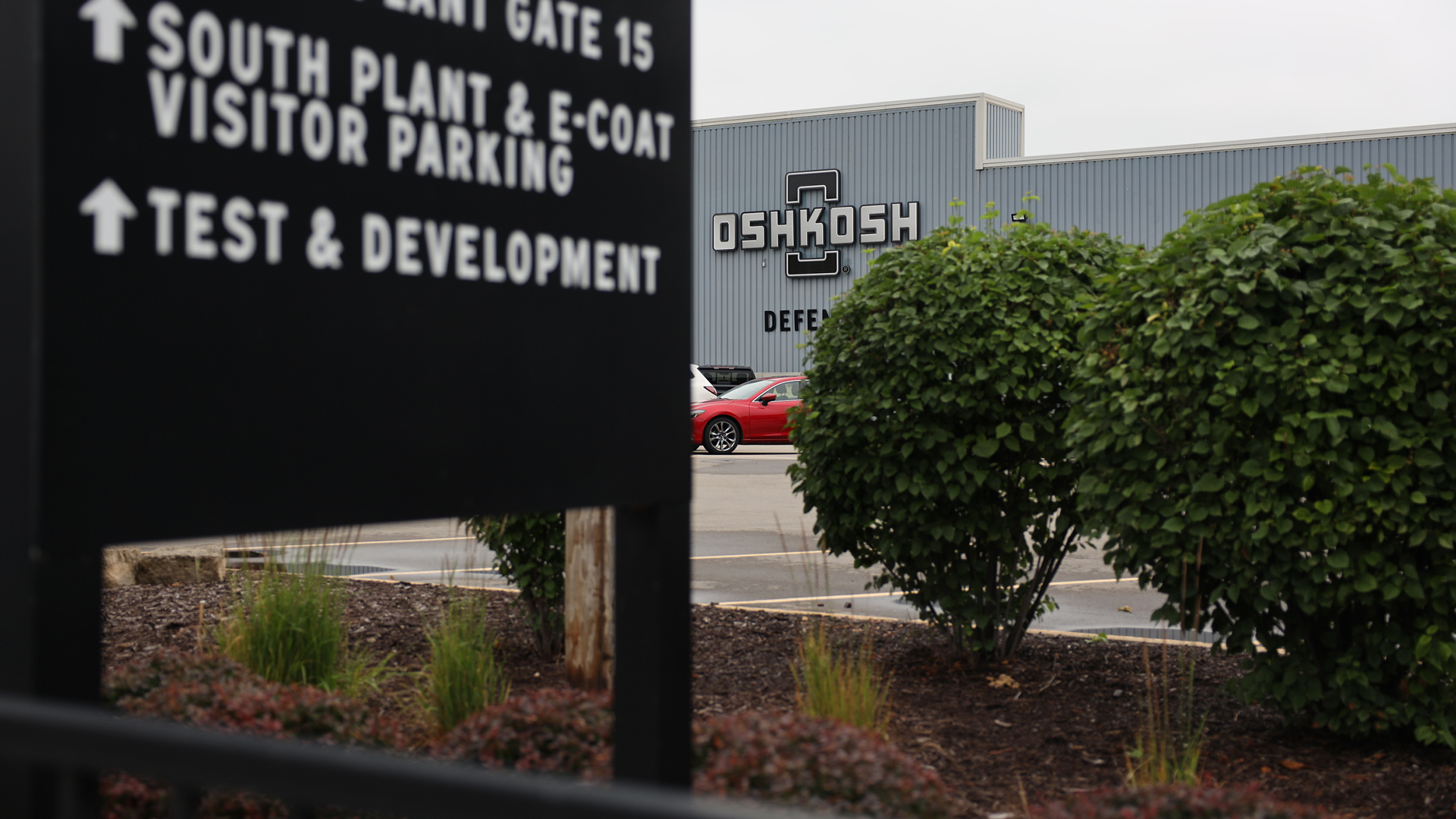 The wordmark for Oshkosh Defense is on the wall of a low-slung building with corrugated metal siding , with bushes and a sign with arrows pointing to "South Plant & E-Coat Visitor Parking" and "Test & Development" in the foreground.