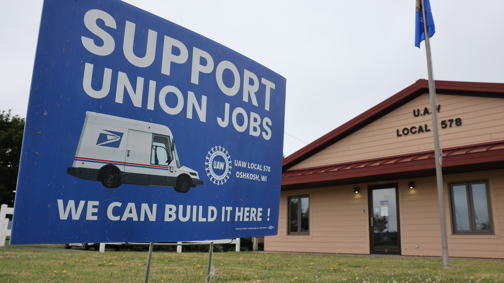 A sign with an illustration of a future postal delivery truck and the slogans "Support Union Jobs" and "We Can Build It Here!" is planted in the lawn in front of a one-story building with a sign reading "U.A.W. Local 578."