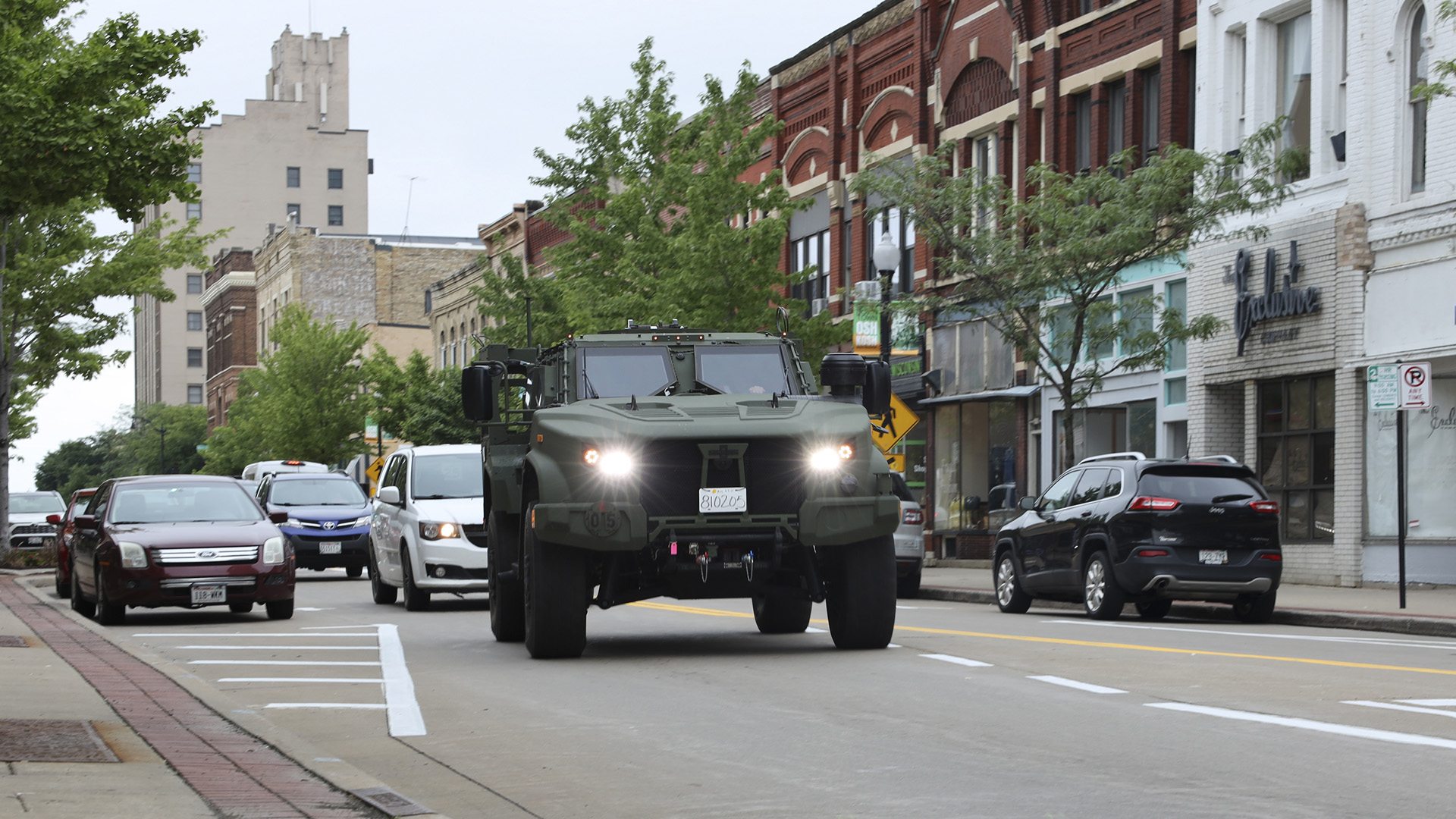 A military vehicle drives down a city street with other vehicles driving and parked and a row of brick storefronts in the background.