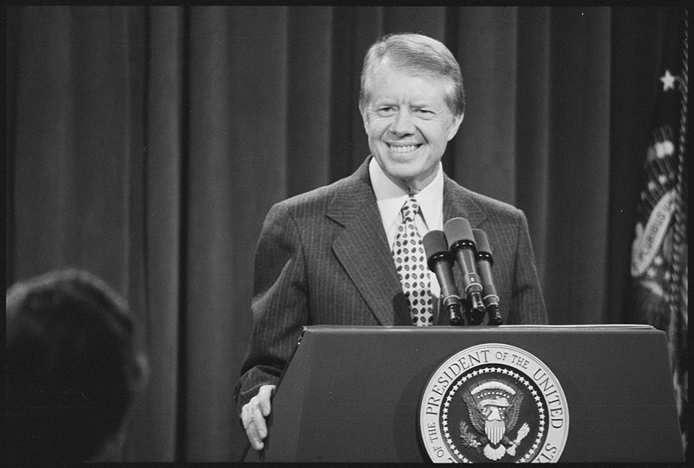 Jimmy Carter stands behind a podium with three microphones and the presidential seal, with a heavy curtain and a flag bearing the presidential seal in the background.