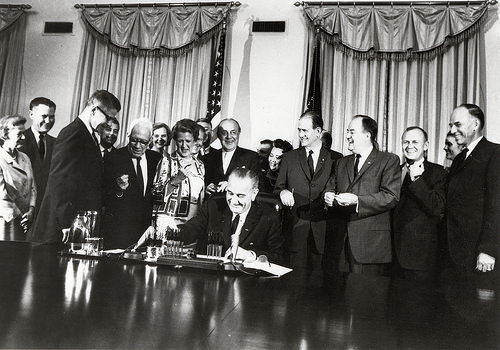 Lyndon Johnson sits at a table and signs a bill into law with more than a dozen people standing in the background.