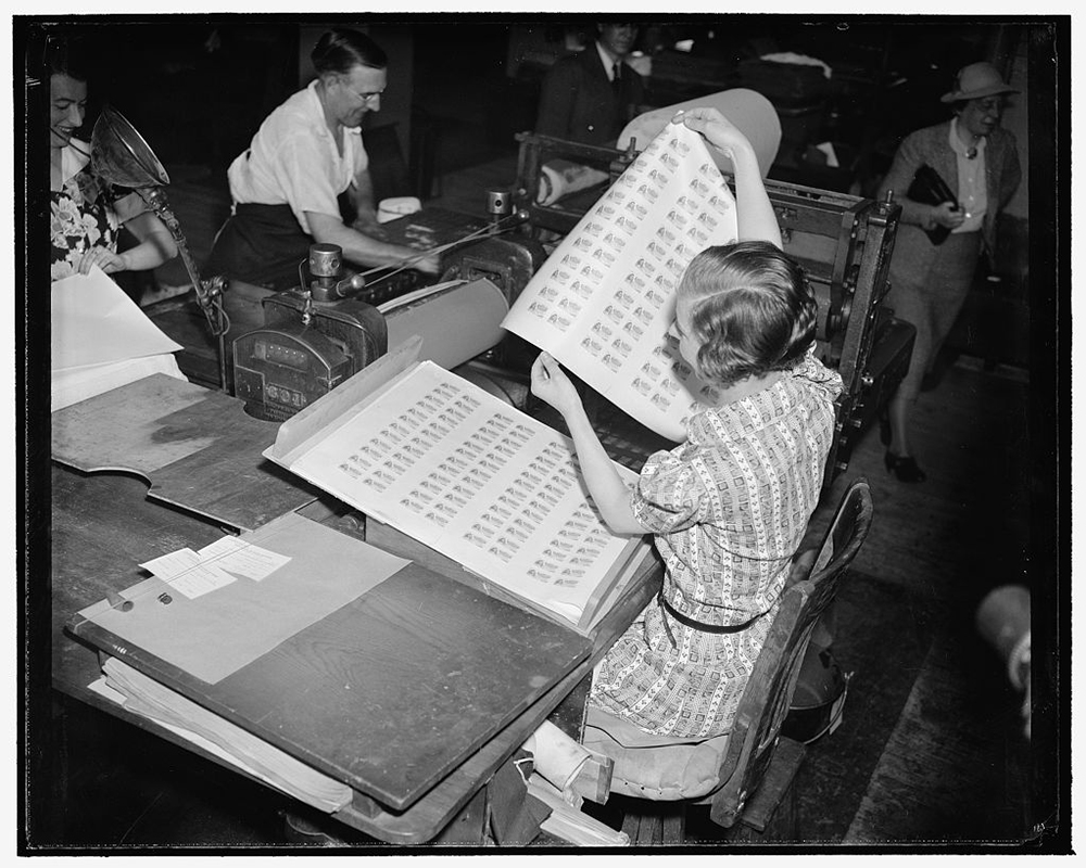 Imogene Stanhope holds a large sheet of paper in the air printed with food stamps, while other workers stand and walk around a printing press.