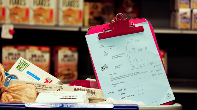 A clipboard holding a checklist with writing sits atop a full grocery cart with boxes of cereal and other food items on shelves in the background.