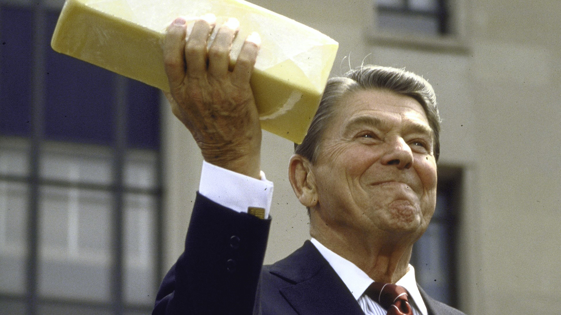 Ronald Reagan holds a large brick-sized piece of cheese in the air with his right hand while smiling.