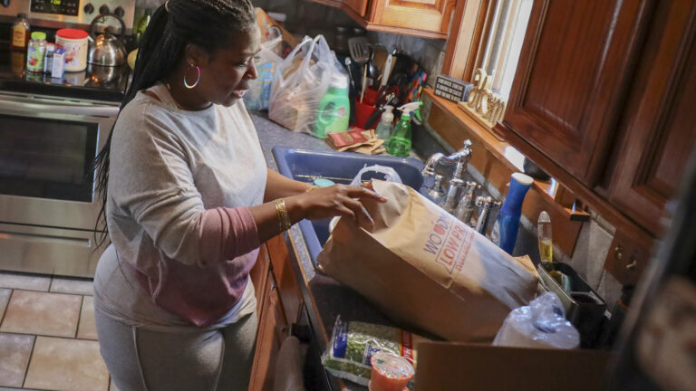 Margaret Benton removes items from a paper grocery bag on a counter with a kitchen sink, oven and more groceries in the background.