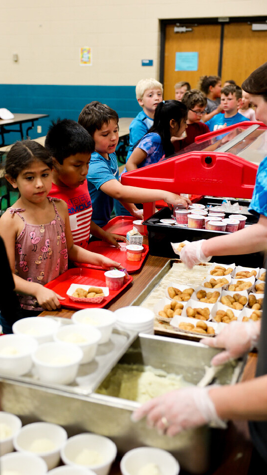 Students stand in line while holding trays and receive items of food inside a school cafeteria.