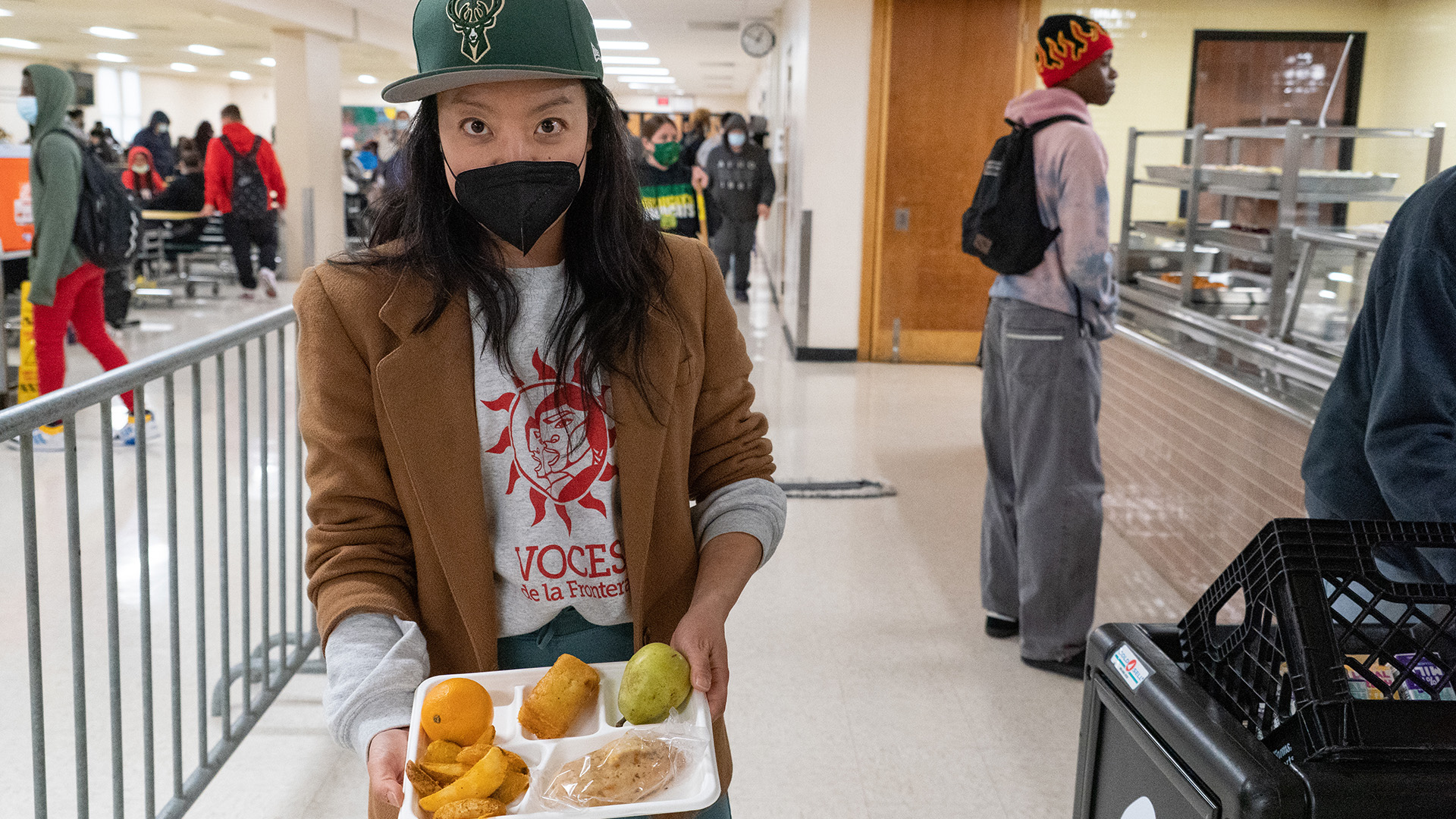 Francesca Hong holds a tray with a school lunch while standing next to a food service counter, with students in a school cafeteria standing and walking in the background.