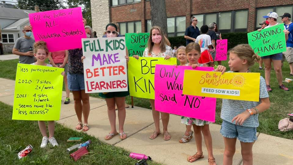 Adults and children stand holding signs with slogans (including "Full Kids Make Better Learners," "Fill the Bellies of All Waukesha Students" and "Starving for Change") while standing on a sidewalk and lawn in front of a school building.