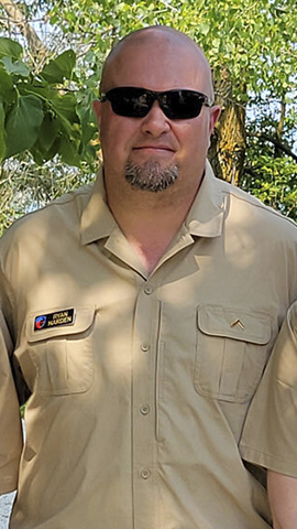 Ryan Harden in a khaki shirt with patches poses for a portrait with trees in the background.