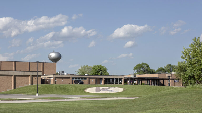 A brick building with one- and two-story sections stands in front of a lawn with a circle K logo on a lawn in the foreground and a water tower with a Kiel label in the background.