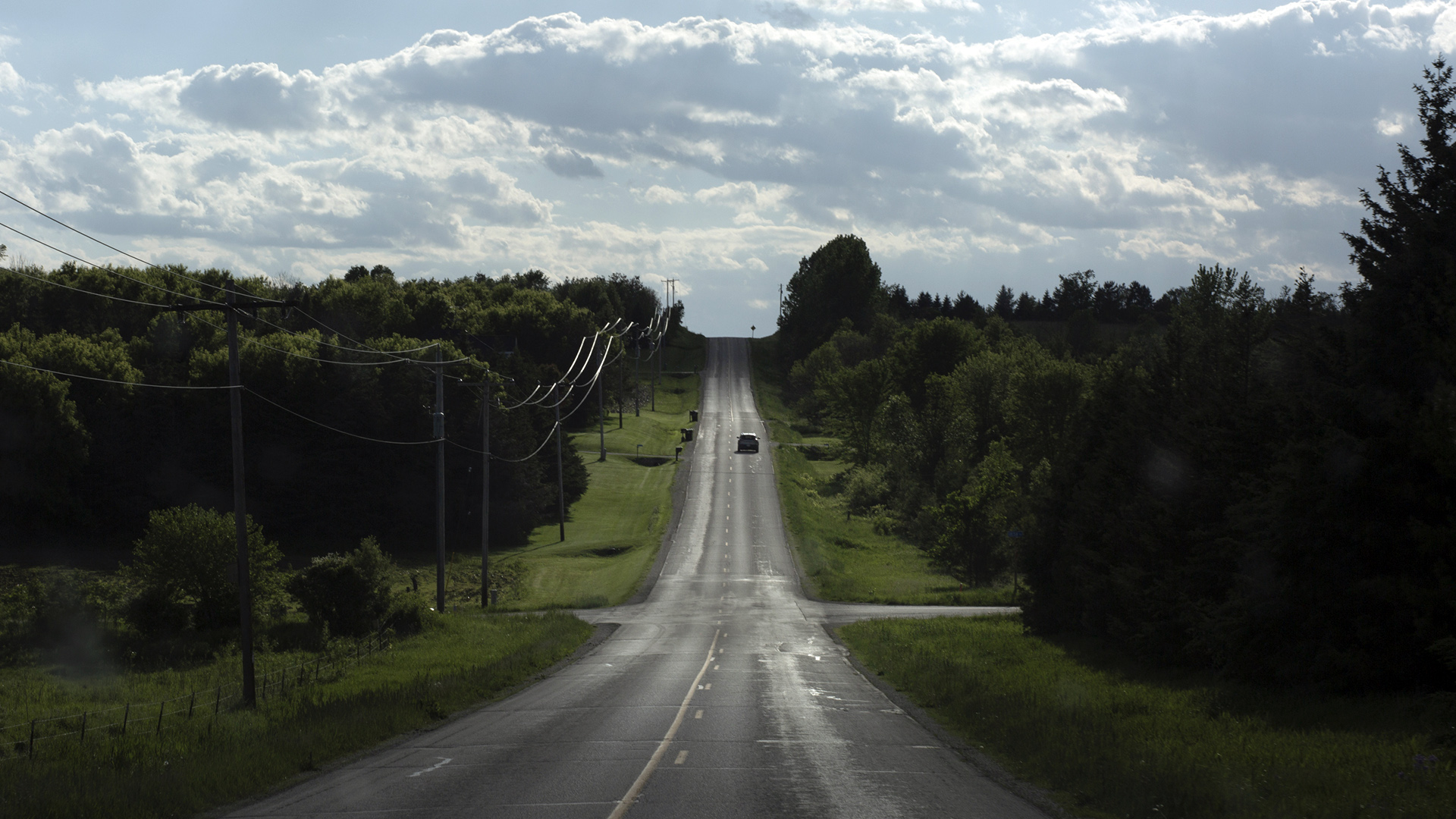 A car drives through a low valley along a two-lane road, with powerlines on one side, trees on both sides, and heavily shadowed clouds in the sky.