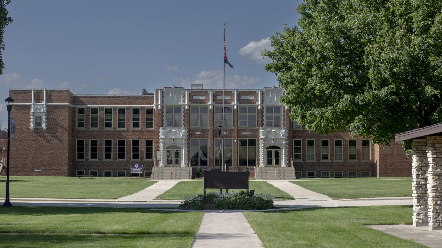 A two-story brick building with a flagpole and sign in front of its doors stands in front of a lawn with a road, sidewalks and trees.