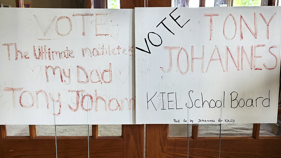 Two handwritten political yard signs read "Vote the Ultimate Mathlete My Dad Tony Johannes" and "Vote Tony Johannes Kiel School Board."