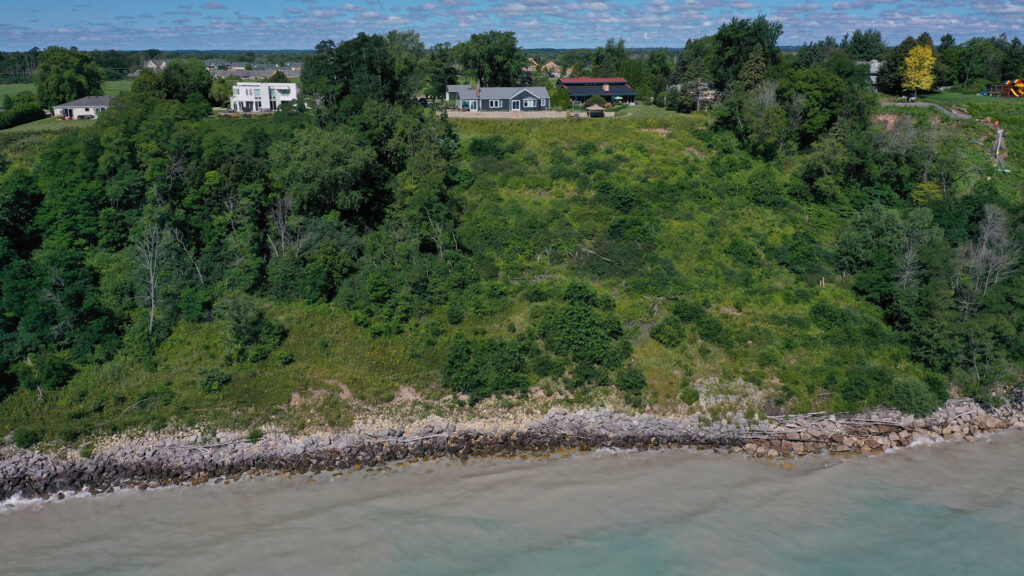 An aerial photo shows a line of houses located near the edge of a bluff covered with trees and other vegetation above a lakeshore comprised of boulders and wood debris.