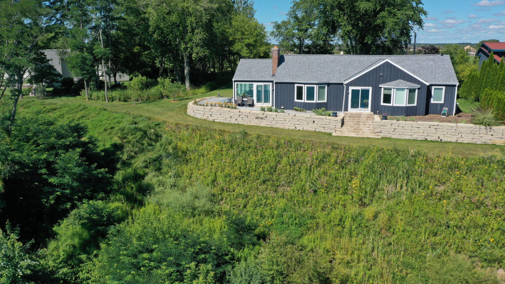 An aerial photo shows a single-story house fronted by a masonry retaining wall in front of a slope covered with various types of vegetation, with trees and other buildings in the background.