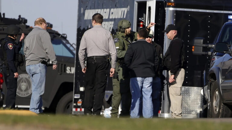Multiple law enforcement officers in uniform, tactical gear and plainclothes stand in front of vehicles and a trailer.