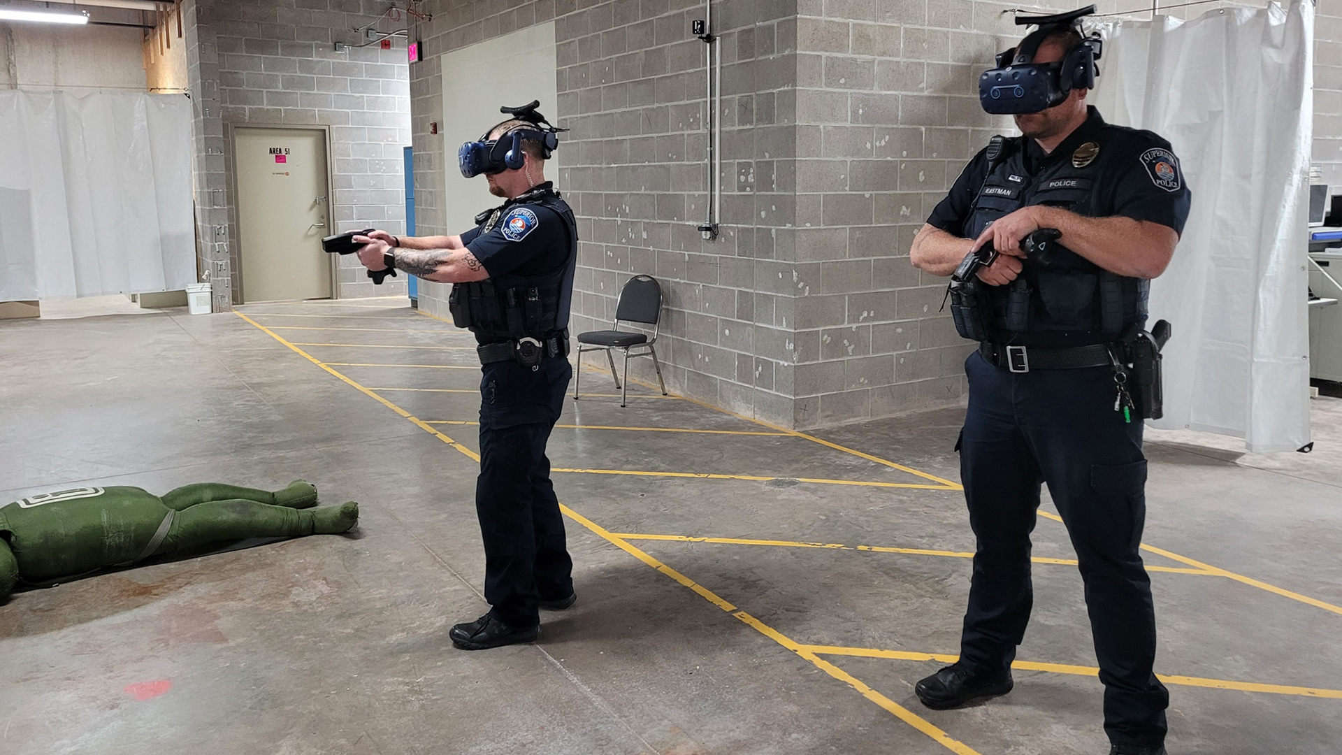 Two police officers standing in a room with concrete block walls and a polished concrete floor with a mannequin lying in a prone position wear virtual reality headsets, with one officer pointing a handgun with two hands and the other holding a handgun in one hand.