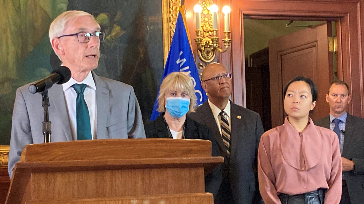Tony Evers stands behind a wooden podium with a microphone in a room with a painting, a wood-paneled wall and door, sconce-style light fixture and Wisconsin flag, with four other people standing to his left in the background.