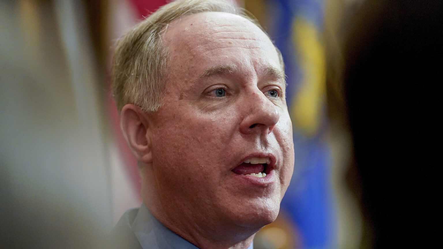 A close-up photo shows Robin Vos speaking with out-of-focus people in the foreground and flags in the background.