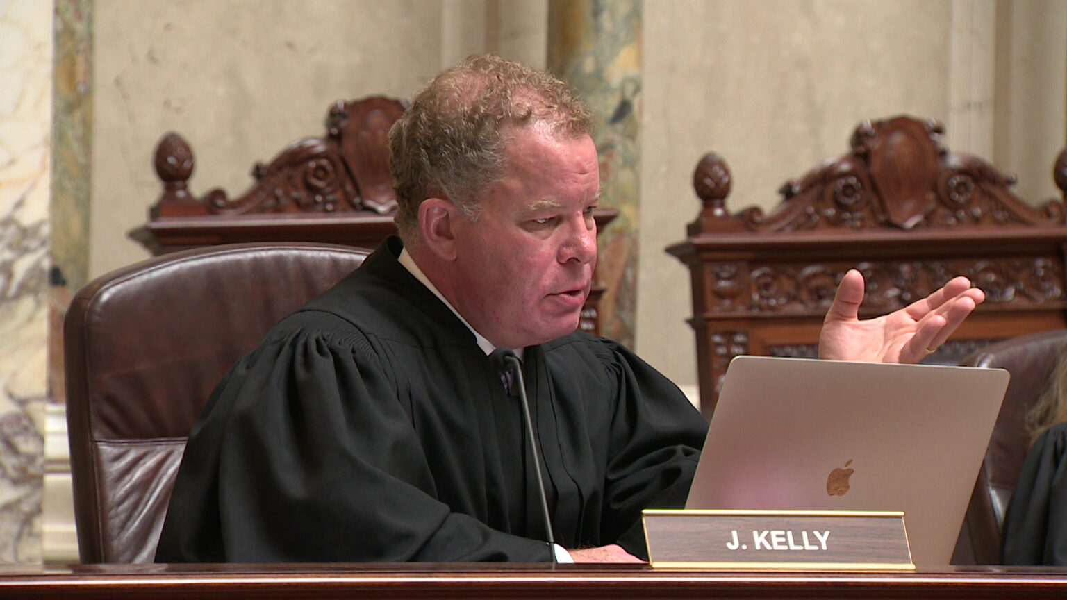 Dan Kelly sits at the justices' bench in the Wisconsin Supreme Court chambers while speaking and gesturing with his left hand while looking at a laptop computer screen.