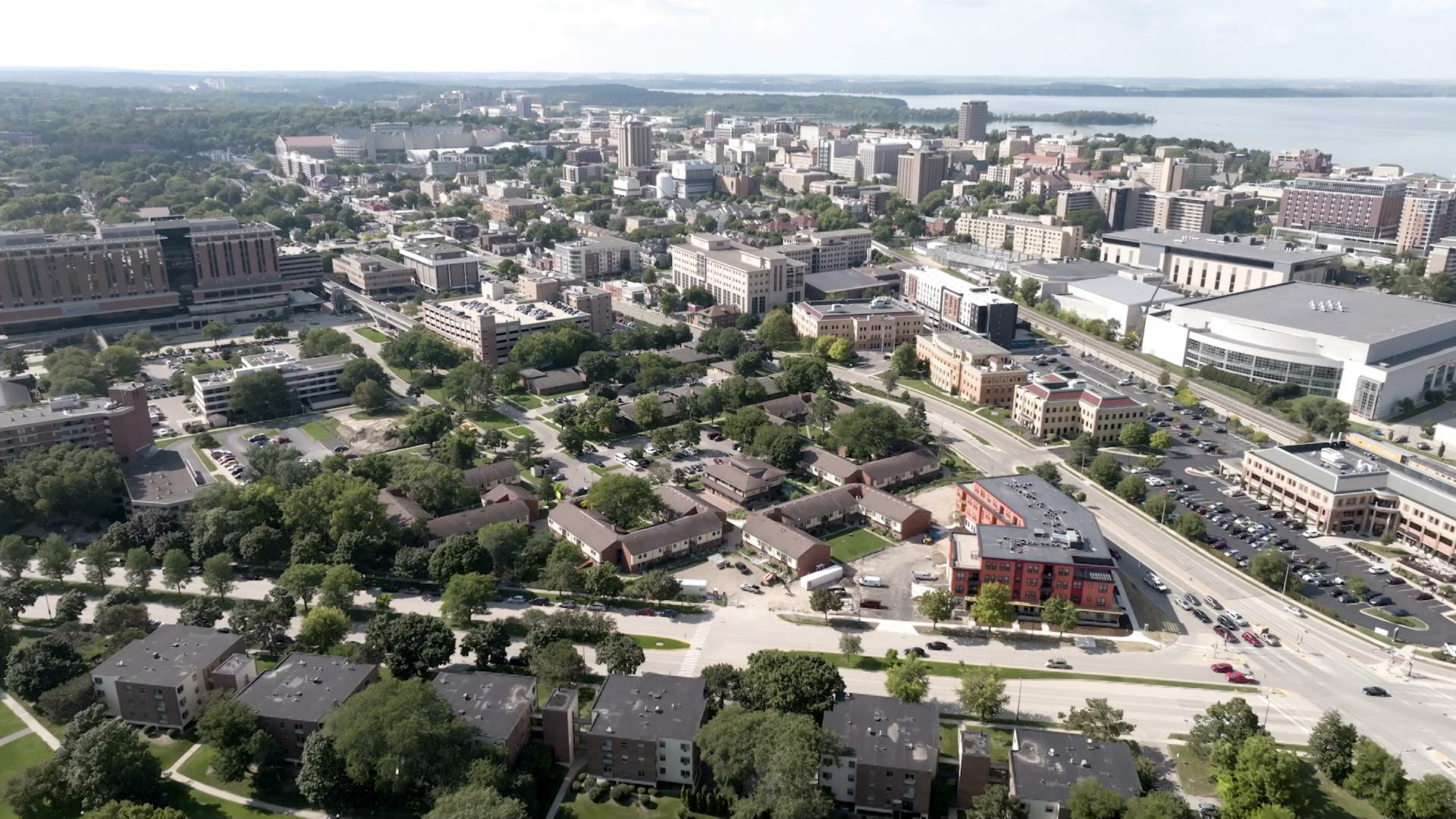 A bird-eye view shows the buildings, roads and trees that make up the streetscape of Madison's Triangle neighborhood and adjacent urbanized areas with large buildings with a portion of a lake in the background.
