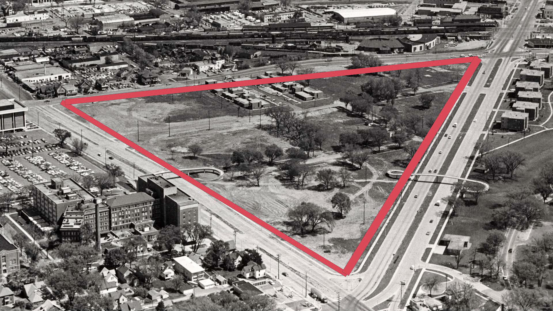 A black-and-white photo shows a red outline along the roads defining an area with trees and mostly empty lots that are surrounded by denser urban development.