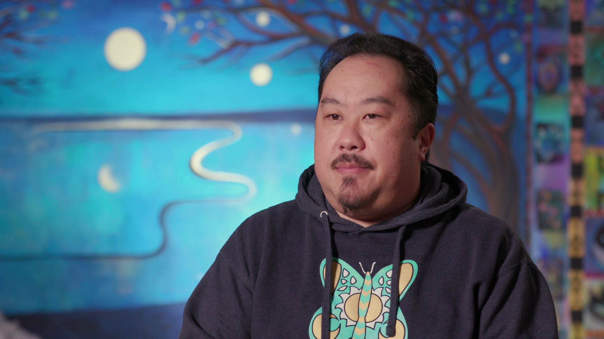 Xong Vang speaks during ah interview with a mural of trees and a night sky in the background.