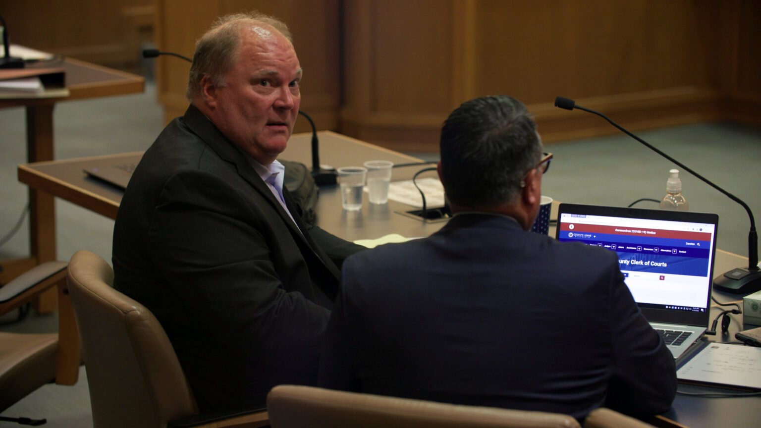 Michael Gableman speaks to another lawyer seated next to him while seated at a counsel table with an open laptop, coffee and water cups, and other items in a courtroom.