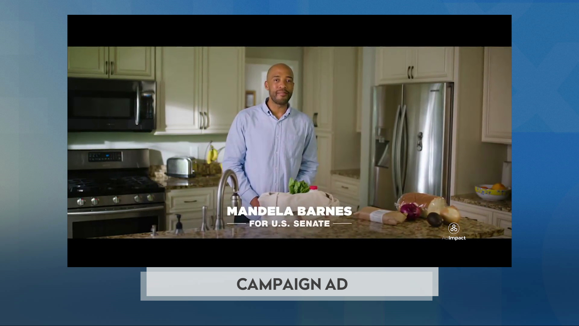A still image from a political advertisement shows Mandel Barnes standing behind a kitchen counter with a sink and grocery items in a bag and sitting on the countertop, with an oven and microwave, refrigerator, painted cupboards and other kitchen items in the background and superimposed with the words "Mandela Barnes For U.S. Senate."