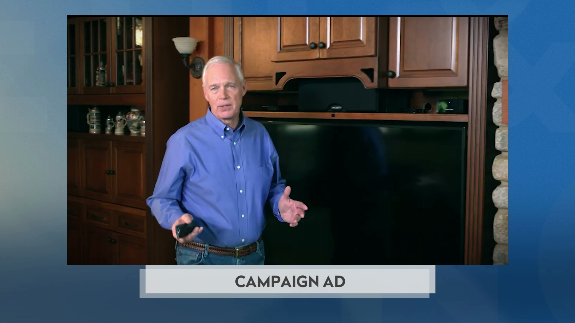 A still image from a political advertisement shows Ron Johnson standing and gesturing with his hands while standing in front of a large wood entertainment center with a television and speakers next to a tall cabinet displaying beer steins.