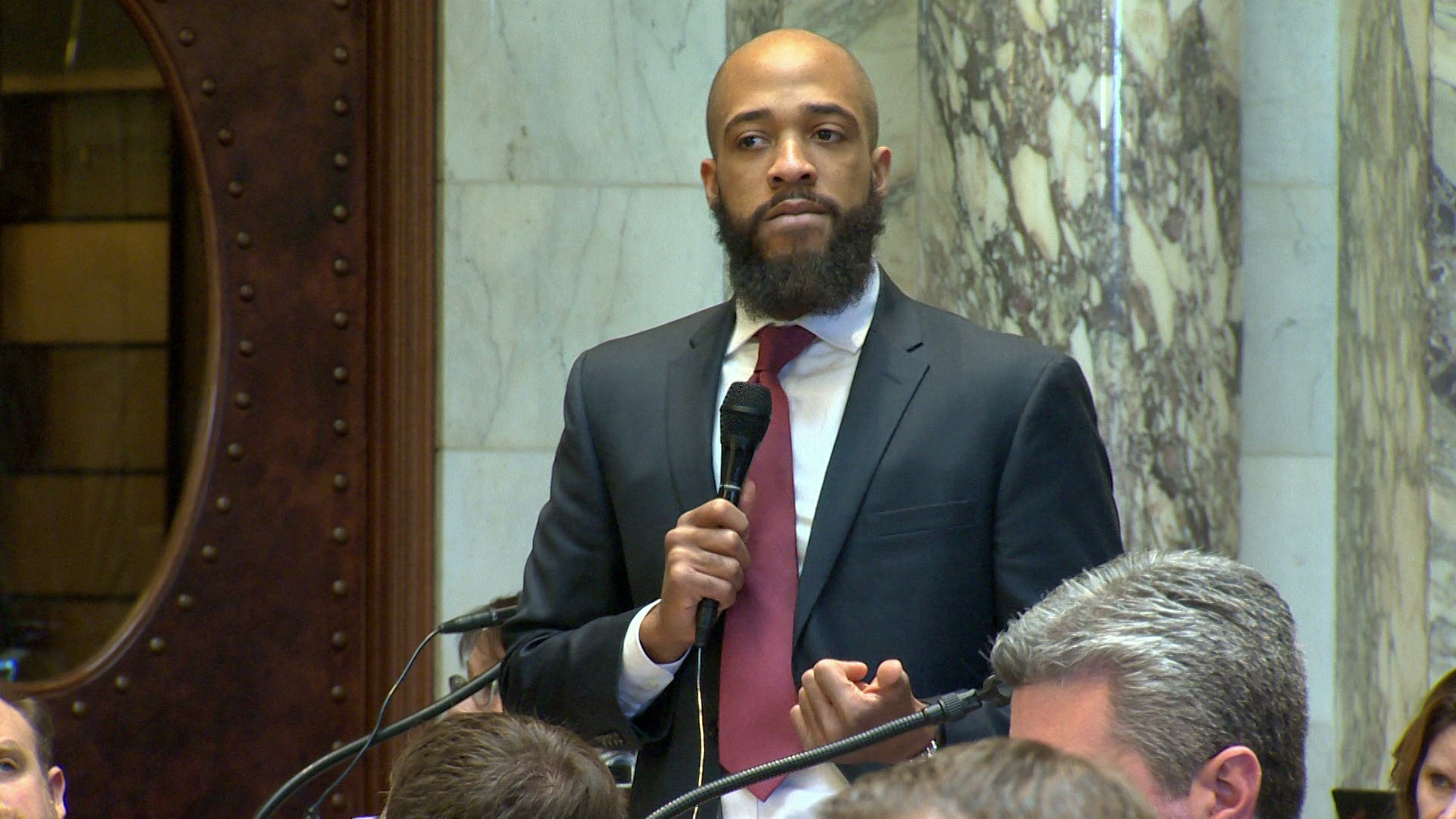 Mandela Barnes speaks into a microphone while standing in a room with marble masonry and pillars, with other people seated around him.