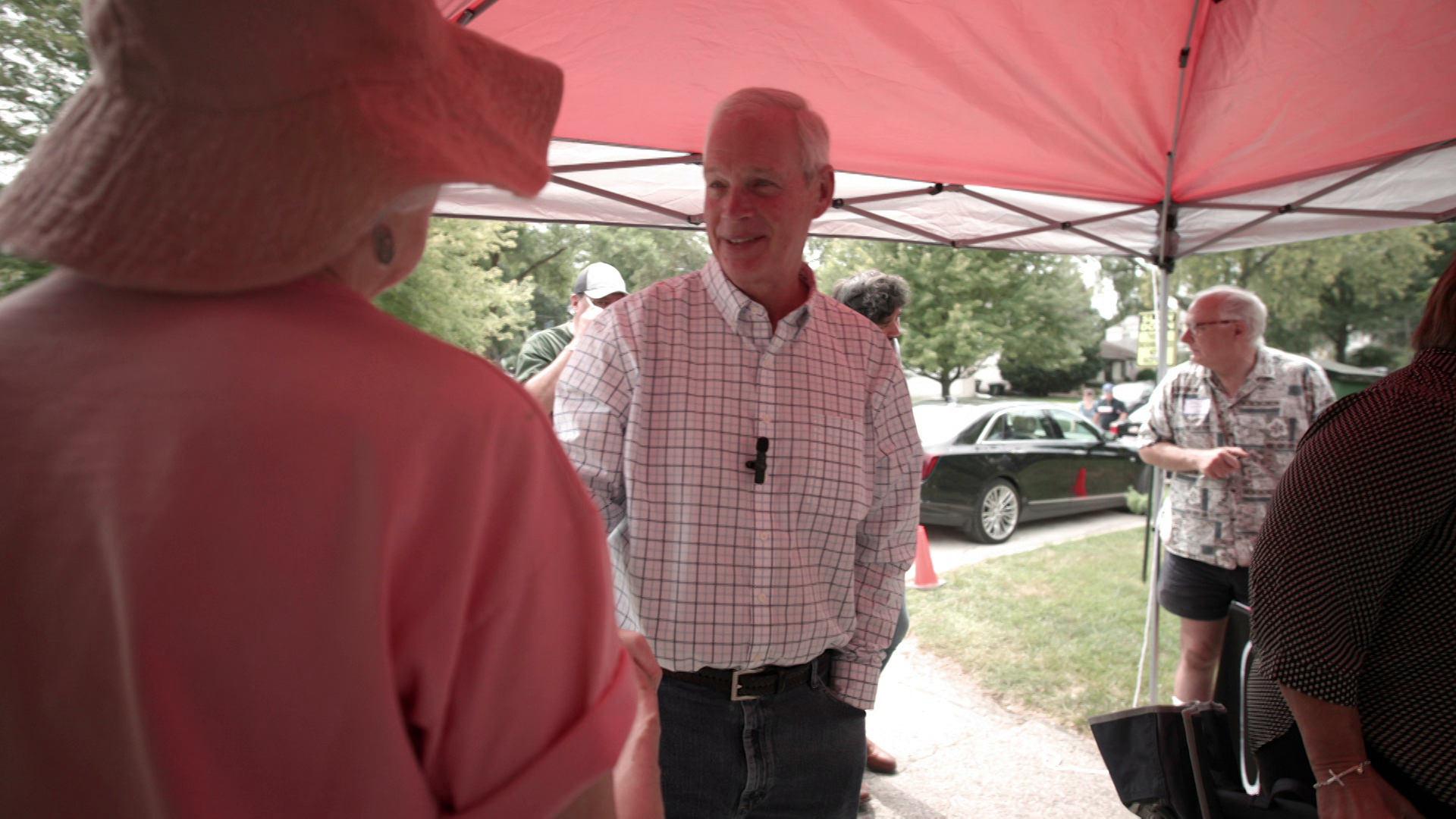 Ron Johnson stands outside and speaks with other people while standing under a canopy tent, with a parked car and trees in the background.