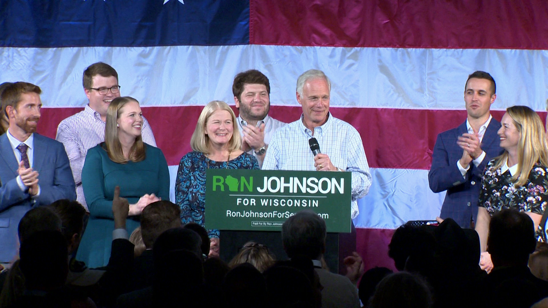 Ron Johnson stands behind a podium with a sign reading "Ron Johnson For Wisconsin" and holds a microphone while speaking, with applauding people standing around him and facing him in the foreground, and with a large U.S. flag as a backdrop in the background.