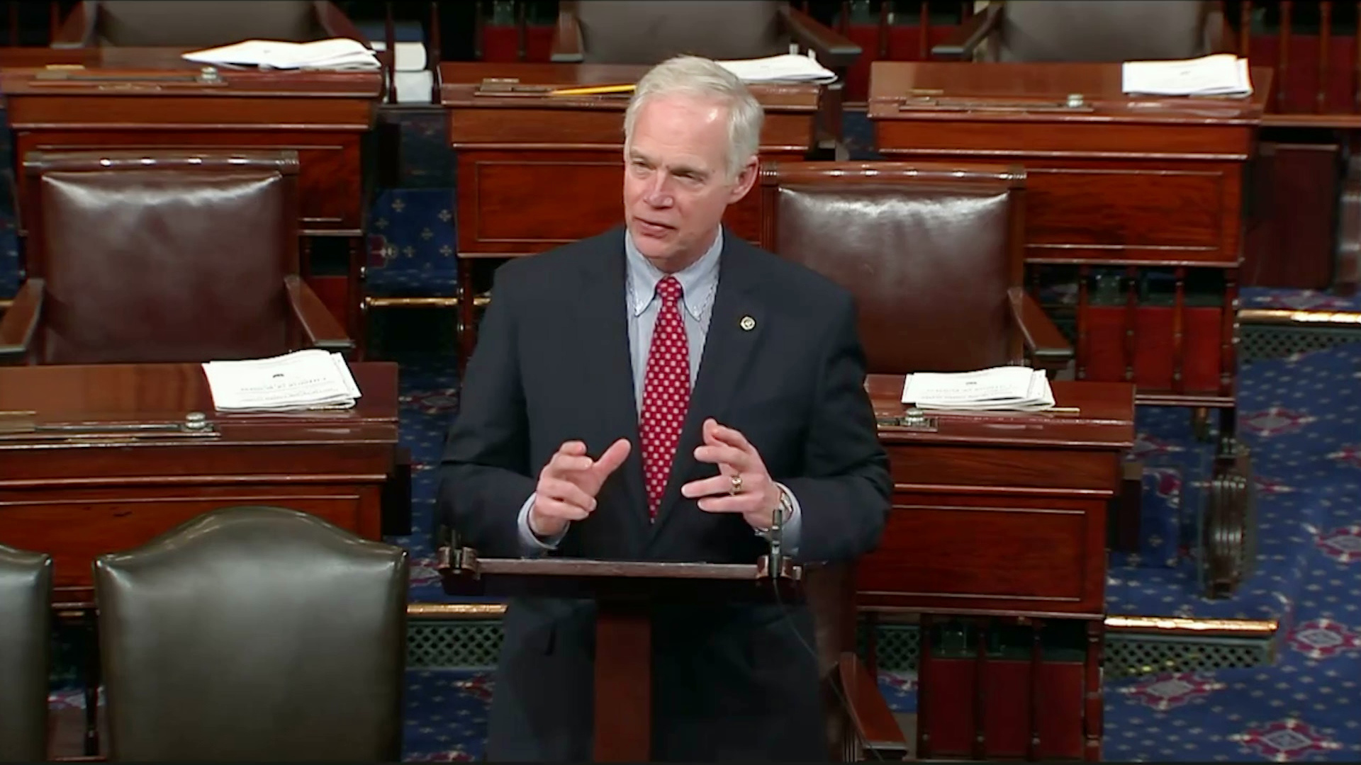 Ron Johnson stands at a wooden podium and speaks while gesturing with his hands, with polished wood desks and leather-padded chairs in the background.