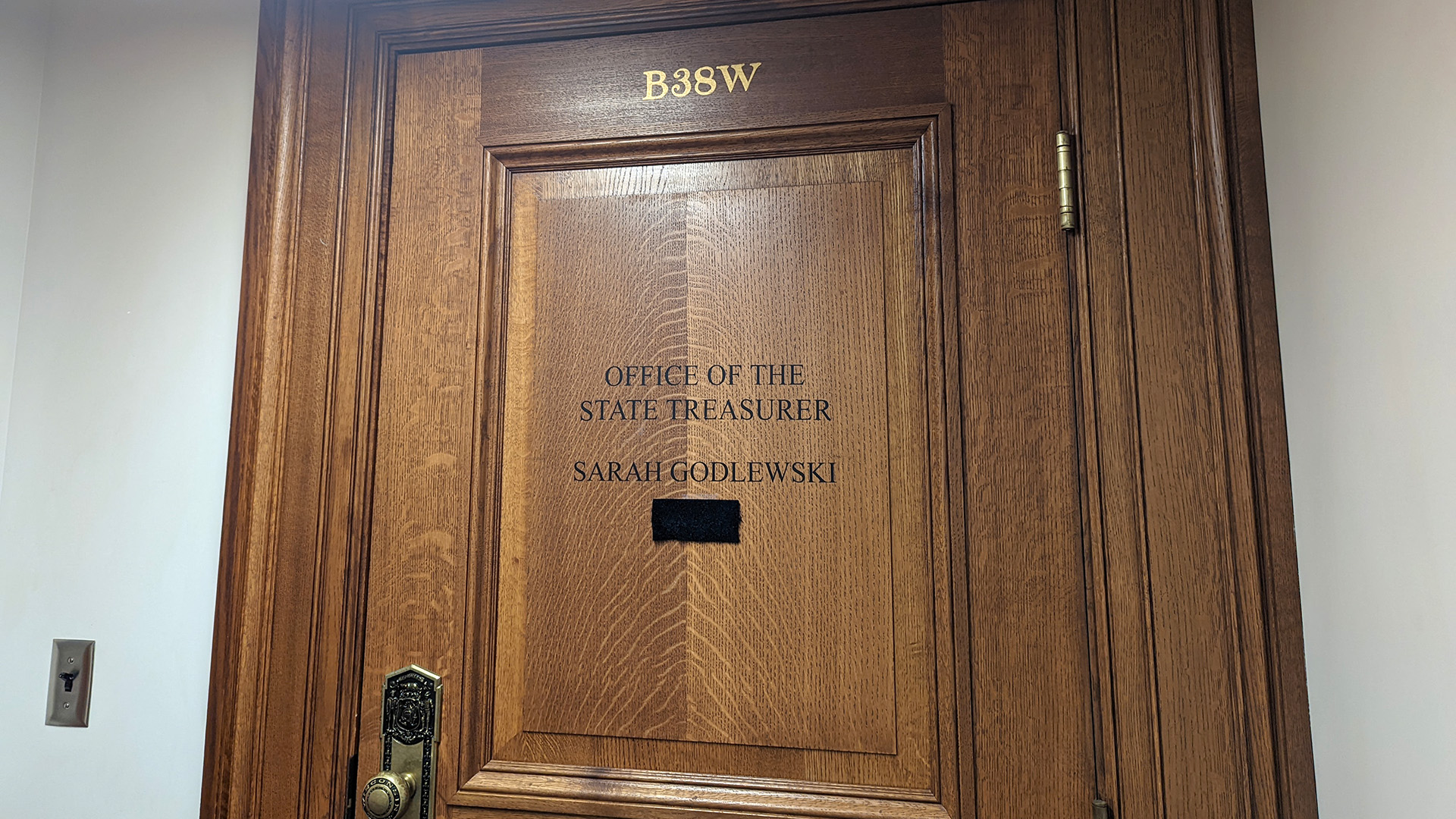 A wood-paneled door with a brass door handle and hinges has the stenciled labels "B38W" and "Office of the State Treasurer Sarah Godlewski" with a velcro patch for attaching items to its surface.