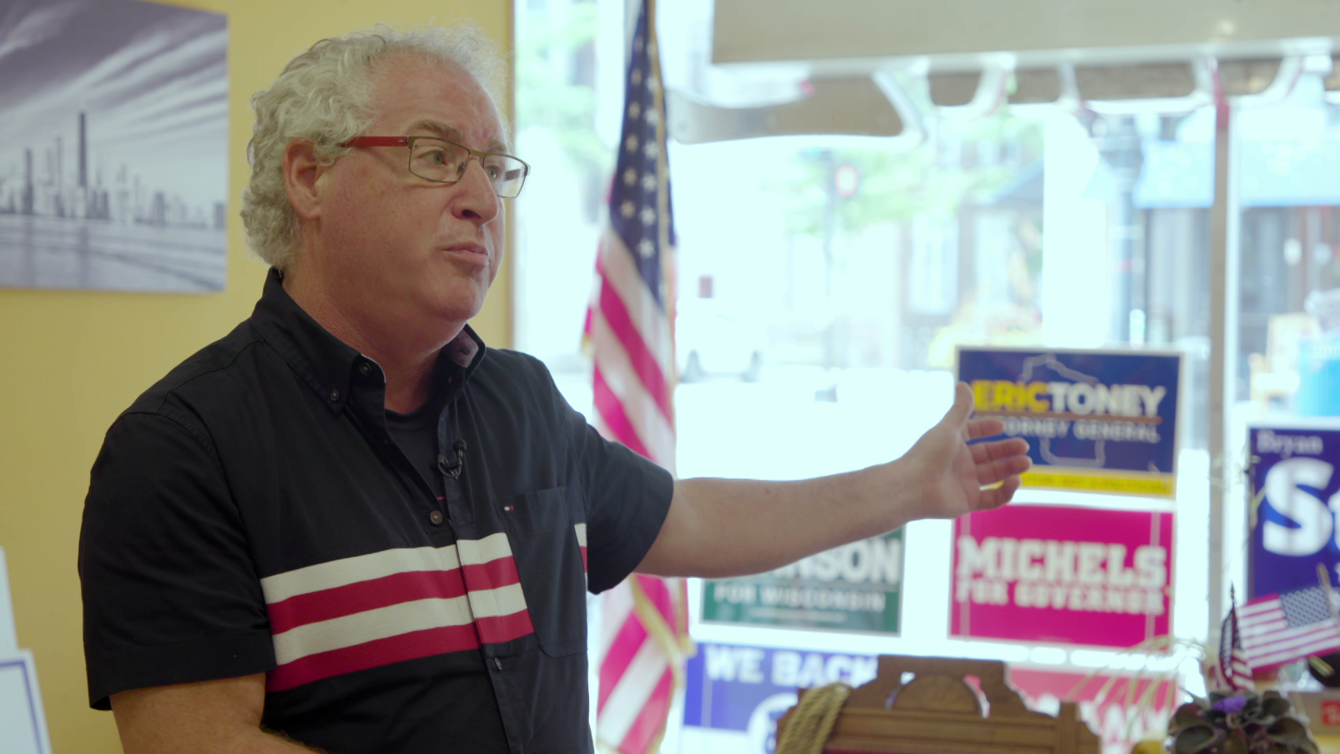 Ken Brown stands in an office and gestures with his left hand with a U.S. flag and political yard signs displayed in a window in the background.