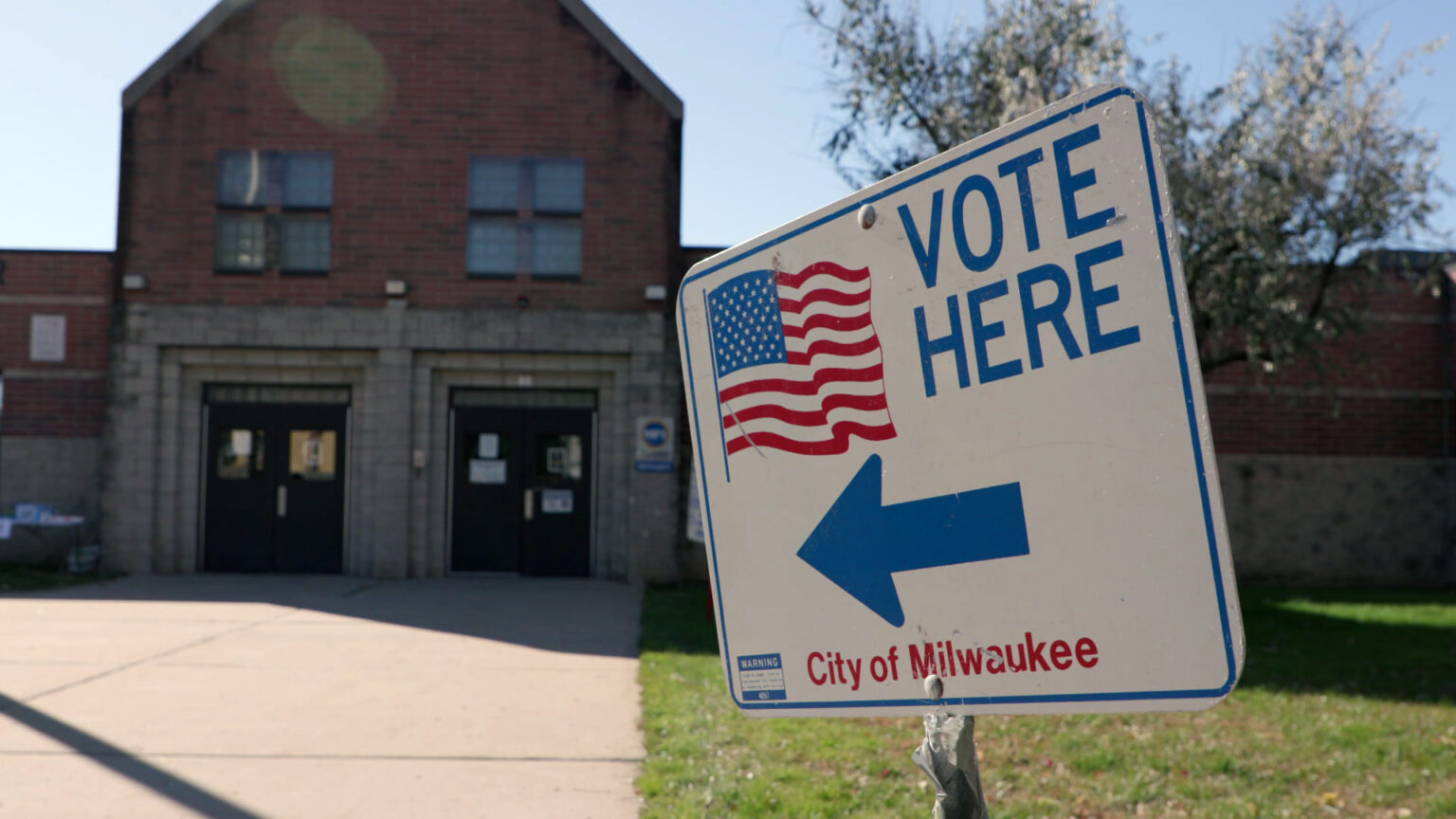 A sign reading Vote Here and City of Milwaukee with a U.S. flag graphic and an arrow is placed in front of a masonry and brick building with two sets of double doors.