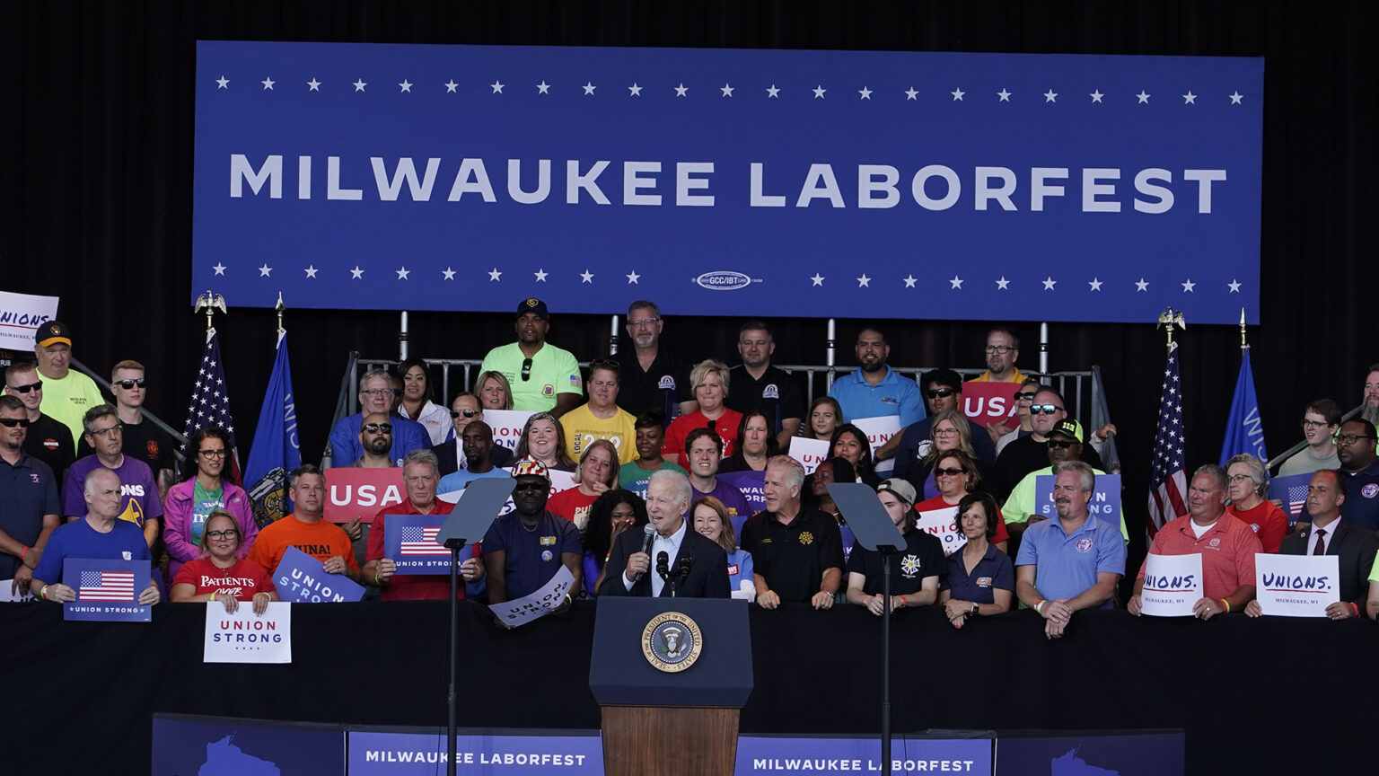 Joe Biden speaks behind a podium with the seal of the President of the United States of America and in front of an audience holding signs that read Union Strong, Unions. and USA with U.S. and Wisconsin flags and a banner reading Milwaukee Laborfest in the background.