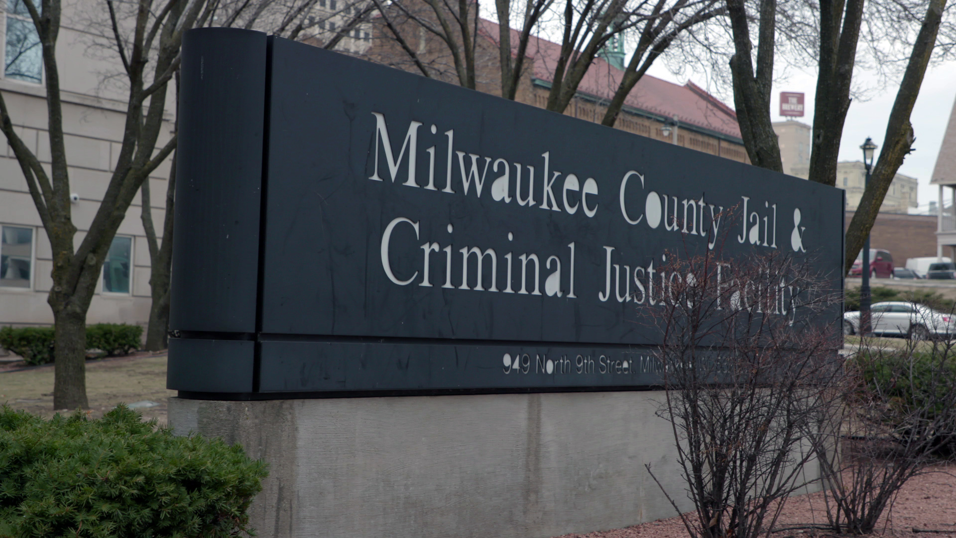 A large sign reading "Milwaukee County Jail & Criminal Justice Facility" with an address reading "949 North 9th Street Milwaukee" stands amid bushes and trees in front of a building, with parked cars and other buildings in the background.
